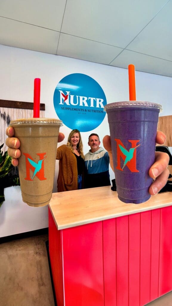 Two colorful smoothies held up in front of a vibrant smoothie bar with smiling staff.